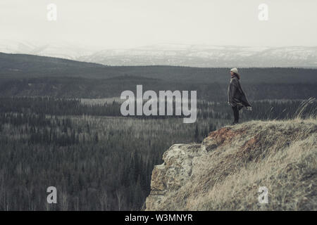 Eine junge Frau steht am Rand einer Klippe mit einer endlosen Blick auf Wald und Berge. Yukon Territory, Kanada Stockfoto