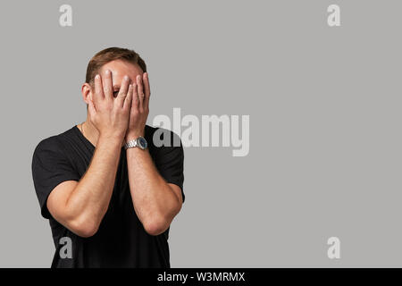 Ein Mann in Freizeitkleidung verbirgt sein Gesicht mit den Händen auf einem grauen Hintergrund. Emotionale, tapferes Gesicht. Auf weissem Hintergrund Platz kopieren Stockfoto