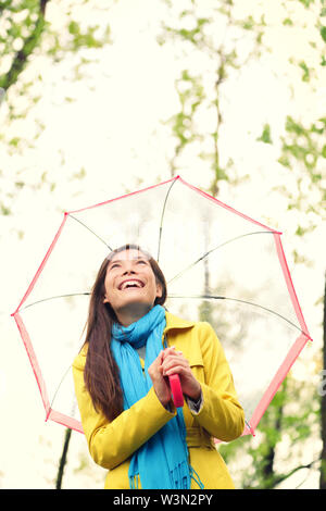 Asiatische Frau im Herbst mit Schirm im Regen freuen. Weibliche Modell zu Clearing Himmel Freude an regnerischen Herbst Tag tragen gelbe Regenmantel in der freien Natur Wald am See. Mischlinge asiatisches Mädchen. Stockfoto