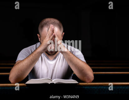 Ein junger Mann sitzt auf einer Kirche Sitzbank, liest in der Bibel und betet. Der Begriff der Religion, Gebet, Glauben Stockfoto