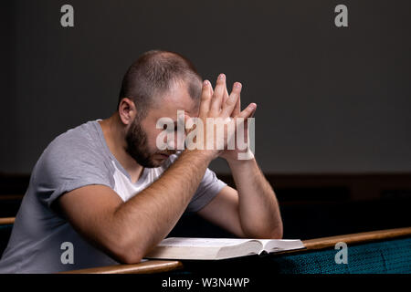 Ein junger Mann sitzt auf einer Kirche Sitzbank, liest in der Bibel und betet. Der Begriff der Religion, Gebet, Glauben Stockfoto