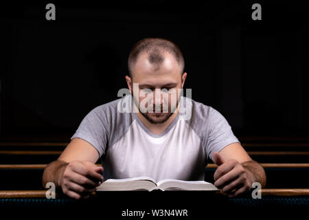 Ein junger Mann sitzt auf einer Kirche Sitzbank, liest in der Bibel und betet. Der Begriff der Religion, Gebet, Glauben Stockfoto