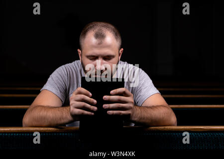 Ein junger Mann sitzt auf einer Kirche Sitzbank, liest in der Bibel und betet. Der Begriff der Religion, Gebet, Glauben Stockfoto