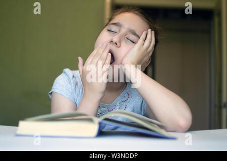 Gefoltert Mädchen liest ein Buch auf dem Tisch und sie ist sehr müde und möchte schlafen, gähnt, schläft beim lesen Geschichten und lehren Stockfoto