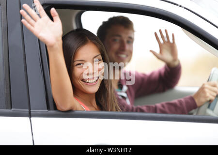 Treiber, die im Auto winken fröhlich in die Kamera. Junges Paar auf Straße Reise im neuen Auto. Interracial glückliches Paar. Stockfoto