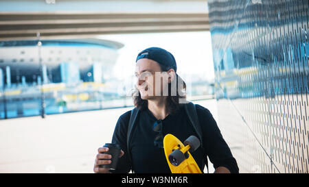 Ein junger Mann in einer Baseball Cap und Denim Shorts und mit einem gelben Skate ist geht von einem metallisch glänzenden reflektierende Wand mit städtischen Ornament. Stockfoto