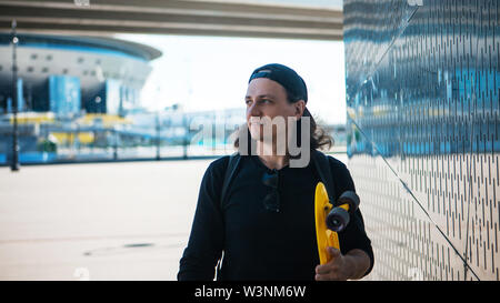 Ein junger Mann in einer Baseball Cap und Denim Shorts und mit einem gelben Skate ist geht von einem metallisch glänzenden reflektierende Wand mit städtischen Ornament. Stockfoto