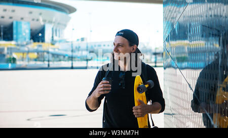 Ein junger Mann in einer Baseball Cap und Denim Shorts und mit einem gelben Skate ist geht von einem metallisch glänzenden reflektierende Wand mit städtischen Ornament. Stockfoto