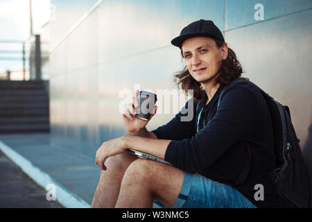 Ein junger Mann in einer Baseball Cap, mit einem Rucksack, und in Denim Shorts, trinkt Kaffee, während er auf den Asphalt, neben einem grauen Granit Wand. Stockfoto