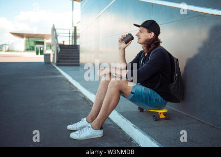 Ein junger Mann in einer Baseball Cap, mit einem Rucksack, und in Denim Shorts, trinkt Kaffee beim Sitzen auf einem gelben Skateboard auf dem Asphalt. Stockfoto