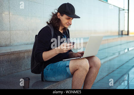 Eine junge dunkelhaarige Mann mit langen Haaren sitzt auf dem Granit Schritte im Schatten mit einem Laptop auf dem Schoß und ein Smartphone. Sommer windigen Tag. Stockfoto