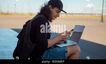Eine junge dunkelhaarige Mann mit langen Haaren sitzt auf dem Granit Schritte im Schatten mit einem Laptop auf dem Schoß und ein Smartphone. Sommer windigen Tag. Stockfoto