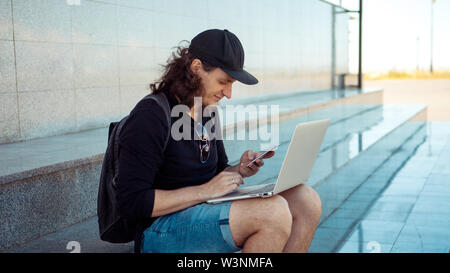 Freelancer arbeiten im Freien, Konzept, jungen Mann mit langen Haaren sitzt auf dem Granit Schritte im Schatten, mit einem Laptop. Sommer windigen Tag. Stockfoto