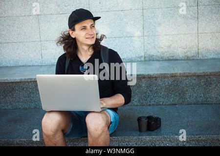 Freelancer arbeiten im Freien, Konzept, jungen Mann mit langen Haaren sitzt auf dem Granit Schritte im Schatten, mit einem Laptop. Sommer windigen Tag. Kopieren Sie sp Stockfoto