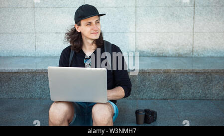 Freelancer arbeiten im Freien, Konzept, jungen Mann mit langen Haaren sitzt auf dem Granit Schritte im Schatten, mit einem Laptop. Sommer windigen Tag. Kopieren Sie sp Stockfoto