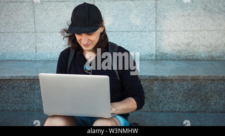 Freelancer arbeiten im Freien, Konzept, jungen Mann mit langen Haaren sitzt auf dem Granit Schritte im Schatten, mit einem Laptop. Sommer windigen Tag. Kopieren Sie sp Stockfoto