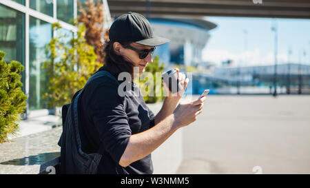 Ein Mann in schwarzen Brille, eine schwarze Baseball Cap und schießt Kaffee auf der Straße auf einem Smartphone. Von der Seite. Paparazzi. Private Ermittlungen. Stockfoto