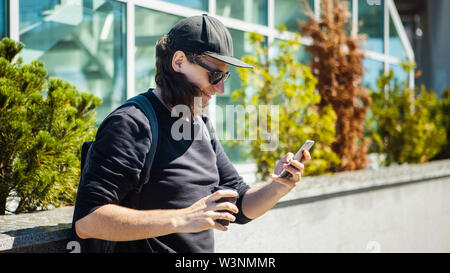 Ein Mann in schwarzen Brille, eine schwarze Baseball Cap und schießt Kaffee auf der Straße auf einem Smartphone. Von der Seite. Paparazzi. Private Ermittlungen. Stockfoto