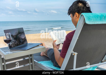 Lesen Buch neben Laptop und einem Glas Wein am Strand Stockfoto