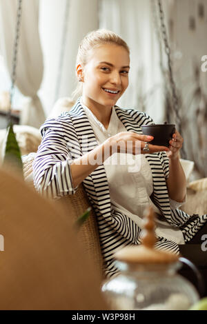 Eingerichtetes Wohnzimmer. Freudige schöne Dame im stilvollen Outfit in ruhiger Atmosphäre beim Trinken frische Tasse Kaffee Stockfoto