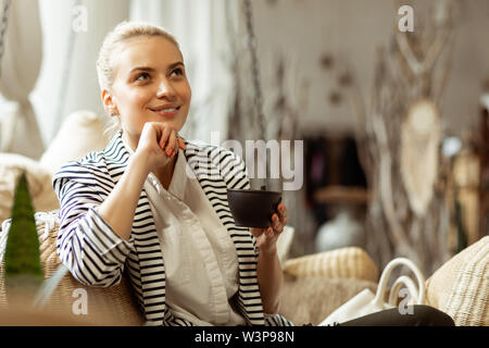 Ruhige Atmosphäre. Nachdenklich gut aussehende Frau mit sanften Lächeln Holding schwarz Tasse Tee beim Chillen im stilvollen Zimmer Stockfoto