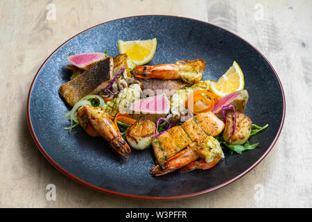 Nahaufnahme der Mix gegrillte Meeresfrüchte und Fisch Platte mit frischem Salat, Garnelen, Jakobsmuscheln, Thunfisch, Lachs, Seebarsch, Zitrone, grüne Soße in einer schwarzen Platte bekennen. Stockfoto