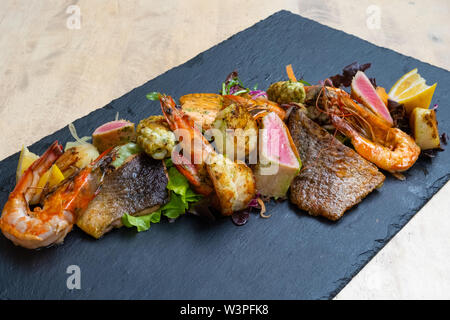 Nahaufnahme der Mix gegrillte Meeresfrüchte und Fisch Platte mit frischem Salat, Garnelen, Jakobsmuscheln, Thunfisch, Lachs, Seebarsch, Zitrone, grüne Soße in einer schwarzen Platte bekennen. Stockfoto