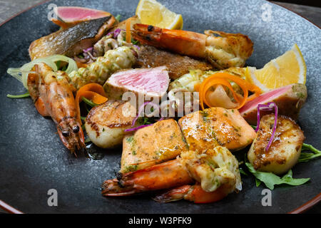 Nahaufnahme der Mix gegrillte Meeresfrüchte und Fisch Platte mit frischem Salat, Garnelen, Jakobsmuscheln, Thunfisch, Lachs, Seebarsch, Zitrone, grüne Soße in einer schwarzen Platte bekennen. Stockfoto