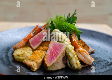 Nahaufnahme der Mix gegrillte Meeresfrüchte und Fisch Platte mit frischem Salat, Garnelen, Jakobsmuscheln, Thunfisch, Lachs, Seebarsch, Zitrone, grüne Soße in einer schwarzen Platte bekennen. Stockfoto