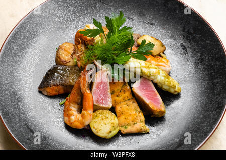 Nahaufnahme der Mix gegrillte Meeresfrüchte und Fisch Platte mit frischem Salat, Garnelen, Jakobsmuscheln, Thunfisch, Lachs, Seebarsch, Zitrone, grüne Soße in einer schwarzen Platte bekennen. Stockfoto