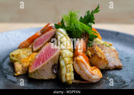 Nahaufnahme der Mix gegrillte Meeresfrüchte und Fisch Platte mit frischem Salat, Garnelen, Jakobsmuscheln, Thunfisch, Lachs, Seebarsch, Zitrone, grüne Soße in einer schwarzen Platte bekennen. Stockfoto