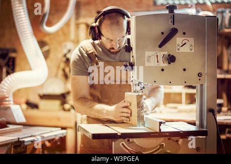 Detail eines Zimmermanns bei der Arbeit schneiden eine Planke mit einer Bandsäge in seiner Werkstatt, Kaukasische mit Bart, eine Schürze und eine Schutzbrille. Stockfoto