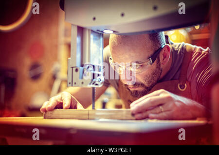 Detail eines Zimmermanns bei der Arbeit schneiden eine Planke mit einer Bandsäge in seiner Werkstatt, Kaukasische mit Bart und Schutzbrille. Stockfoto