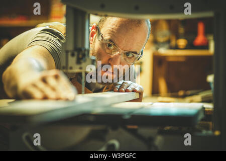 Detail eines Zimmermanns bei der Arbeit schneiden eine Planke mit einer Bandsäge in seiner Werkstatt, Kaukasische mit Bart und Schutzbrille. Stockfoto