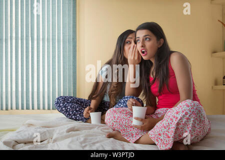 Zwei junge Mädchen sitzen auf dem Bett zu Hause halten Kaffeetassen und klatschen Stockfoto