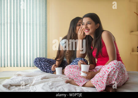 Zwei lächelnde junge Mädchen sitzen auf dem Bett zu Hause halten Kaffeetassen und klatschen Stockfoto