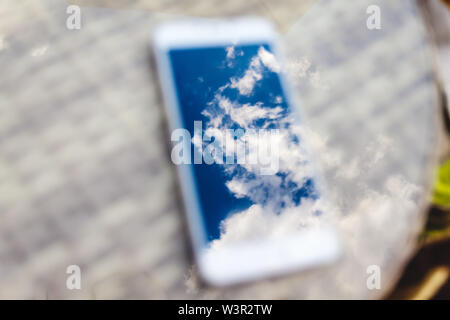Reflexion der blauen Himmel mit Wolken im Telefon - Smartphone, das auf einem hellen Rattanmöbeln Tabelle vor dem Hintergrund liegt. Blur und Abstraktion Stockfoto
