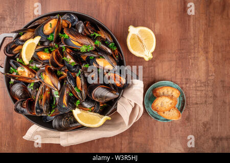 Marinara Muscheln, Moules Mariniere, mit geröstetem Brot und Zitronenscheiben, im Kochtopf, Overhead shot auf einem dunklen Holzmöbeln im Landhausstil Hintergrund Stockfoto