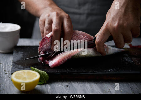 Nahaufnahme eines jungen kaukasischen Mann Schneiden eine rohe frische Makrelen mit einem Messer, auf einer Schiefertafel Fach auf einem rustikalen hölzernen Tisch oder Arbeitsplatte platziert Stockfoto