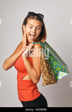 Junge brünette Dame im roten T-Shirt mit Sonnenbrille mit Tüten posing und Kamera über weißen Hintergrund schockiert. Stockfoto