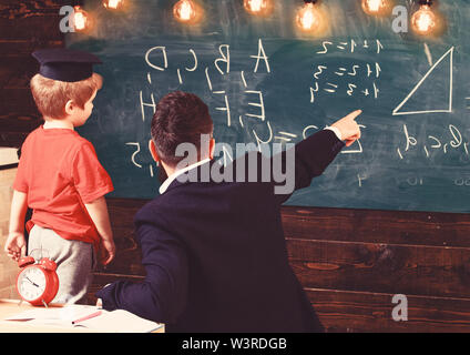 Junge männliche Lehrer führt sein Kind Schüler lernen während zeigen und auf Schiefertafel mit kritzelt auf, sitzen im Klassenzimmer, Ansicht von hinten. Stockfoto