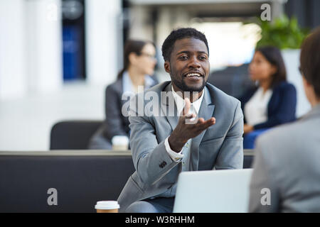 Positive zuversichtlich gutaussehenden jungen Schwarzen bärtigen Geschäftsmann in grauen Anzug in der Lobby sitzen und Gestik, die Hand beim Gespräch mit dem Geschäftspartner während Stockfoto