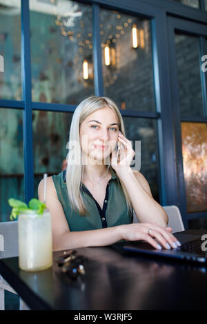 Eine junge schöne blonde Mädchen sitzt in einem gemütlichen Café und arbeitet für einen Laptop, Getränke ein erfrischendes Getränk. Rest in der Hitze des Sommers Stockfoto