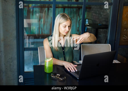 Eine junge schöne blonde Mädchen sitzt in einem gemütlichen Café und arbeitet für einen Laptop, Getränke ein erfrischendes Getränk. Rest in der Hitze des Sommers Stockfoto