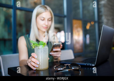 Eine junge schöne blonde Mädchen sitzt in einem gemütlichen Café und arbeitet für einen Laptop, Getränke ein erfrischendes Getränk. Rest in der Hitze des Sommers Stockfoto