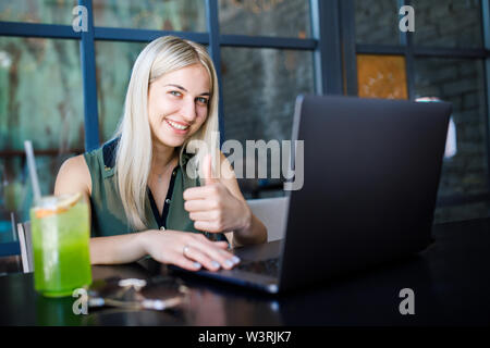 Eine junge schöne blonde Mädchen sitzt in einem gemütlichen Café und arbeitet für einen Laptop, Getränke ein erfrischendes Getränk. Rest in der Hitze des Sommers Stockfoto