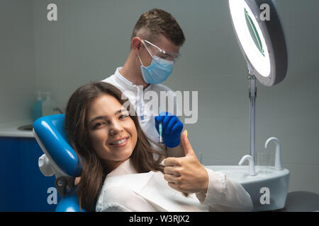Schöne junge lächelnde brünette Frau, die Untersuchung bei einer Zahnarztpraxis durch stattliche Zahnarzt in Maske und blaue Handschuhe. Stockfoto