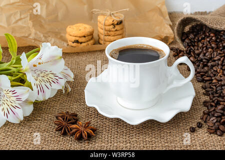 Weiß peruanischen Lilie Blume, heißen Kaffee in Weiß elegante Tasse mit Untertasse, süße Kekse und Streuung von Kaffeebohnen auf rustikalen Sack Hintergrund. Stockfoto