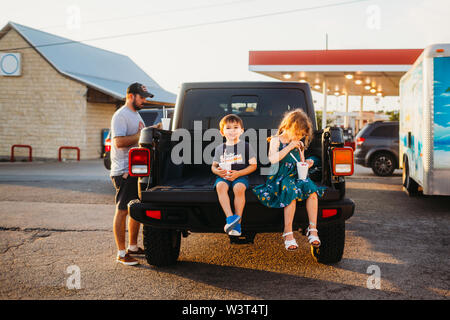 Jungen und Mädchen sitzen auf jeep Truck Bed essen Schnee Kegel mit Vati Stockfoto