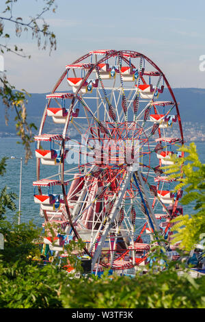 Einen bunten Riesenrad im Vergnügungspark am sonnigen Sommertag über blauen Himmel Hintergrund. Vorderansicht durch grüne Bäume. Unterhaltung anhand von quantitativen Simulatio Stockfoto